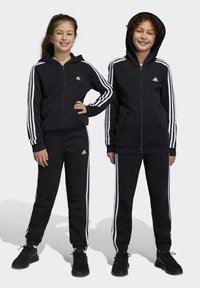 Sudaderas con capucha negras con cremalleras y acentos de tres rayas blancas en las mangas y los laterales, combinadas con joggers negros a juego.