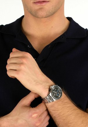 Mann in schwarzem Polo passt eine silberne Metallarmbanduhr mit schwarzem Zifferblatt an seinem linken Handgelenk vor weißem Hintergrund an.