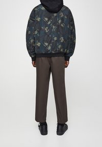 Kurtka bombowca w kamuflażu w kolorze zielonym i czarnym, z prążkowanymi mankietami, noszona na brązowych spodniach i czarnych sneakersach, prezentująca luźny krój i nowoczesny design.