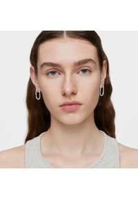 Orecchini a cerchio in argento dalla forma ovale, con una superficie liscia e lucida e un design minimalista. Indossati da una modella con i capelli lunghi.