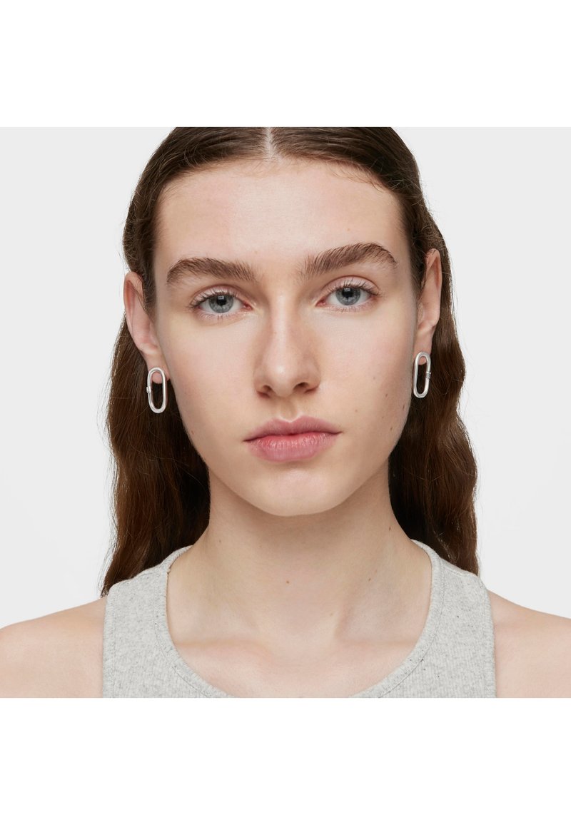 Orecchini a cerchio in argento dalla forma ovale, con una superficie liscia e lucida e un design minimalista. Indossati da una modella con i capelli lunghi.