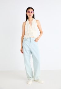 Top blanco de halter con un dobladillo ondulado, combinado con vaqueros de tiro alto azul claro y zapatillas blancas. La modelo lleva gafas de sol rosas.