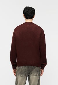 Homme portant un pull en maille rouge foncé et un jean gris délavé, vu de dos devant un fond clair uni.