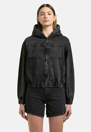 Kvinde iført sort denim-hættetrøje med frontlommer og matchende sorte denimshorts, stående mod en ensfarvet lys baggrund.