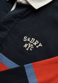 Donkerblauwe polo met een witte kraag, voorzien van een geborduurd "S&DRY NYC" logo in crèmekleur, en meerkleurige strepen in rood, blauw en oranje.