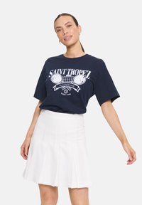 T-shirt bleu marine avec le texte graphique blanc "SAINT TROPEZ" et des raquettes de tennis, associé à une mini-jupe plissée blanche. Texture de tissu lisse.