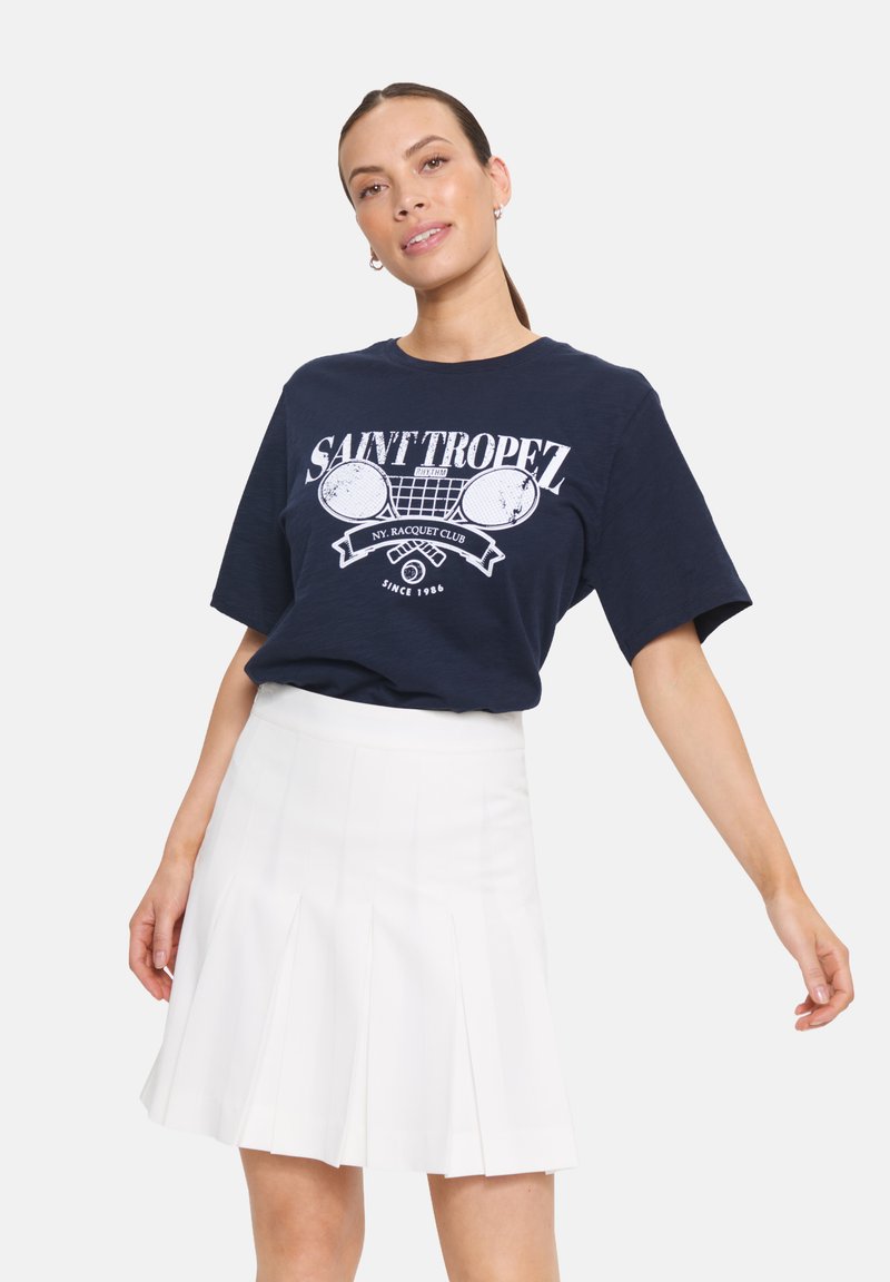 T-shirt bleu marine avec le texte graphique blanc "SAINT TROPEZ" et des raquettes de tennis, associé à une mini-jupe plissée blanche. Texture de tissu lisse.