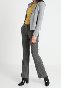Blazer gris con un corte entallado, combinado con una blusa de color mostaza. Pantalones grises texturizados y botas de tacón negro brillante completan el conjunto.