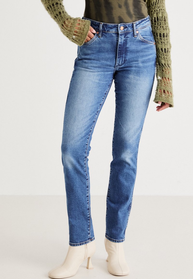 Jeans de mezclilla azul de tiro alto con un acabado desgastado, cinco bolsillos y pasadores para cinturón, combinados con un suéter de punto verde y botas de tobillo beige.