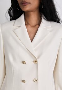 White blazer with a notched collar, featuring gold square-shaped buttons and a structured silhouette, crafted from a smooth fabric.