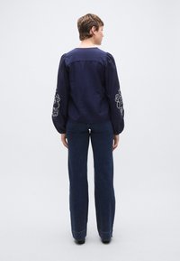 Μπλούζα σε ναυτικό μπλε με υφασμάτινη υφή, με φουσκωτά μανίκια διακοσμημένα με λευκή κεντητή ανθοδέσμη, σε συνδυασμό με σκούρα μπλε τζιν παντελόνια με φαρδιά μπάρα.