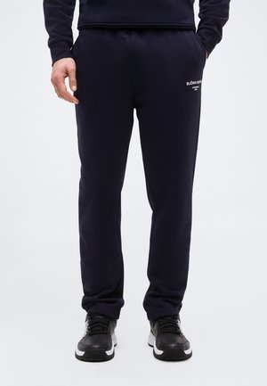 Άτομο που φοράει μαύρο παντελόνι φόρμας Björn Borg και μαύρα αθλητικά παπούτσια, στέκεται με το ένα χέρι στην τσέπη μπροστά σε απλό, φωτεινό φόντο.