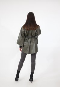 Grüne Jacke aus Kunstleder mit tailliertem Schnitt, weiten Ärmeln und lockerem Sitz, kombiniert mit grauen Leggings und schwarzen Stiefeln mit hohem Absatz.