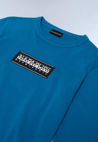 Kék pamuts póló kerek nyakkal és rövid ujjakkal, a mellkasán egy téglalap alakú fekete-fehér "NAPAPURDI geographic" logóval.