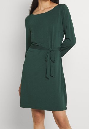 Femme portant une robe vert foncé longueur genou avec manches longues et ceinture en tissu nouée à la taille sur un fond clair uni.