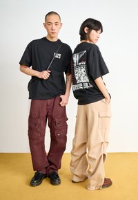 Two models wear casual streetwear. One in a black graphic t-shirt and burgundy cargo pants; the other in a black graphic t-shirt and beige pants.