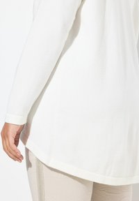 Top bianco a maniche lunghe con scollatura rotonda e texture liscia, caratterizzato da spacchetti laterali e orlo posteriore leggermente allungato.