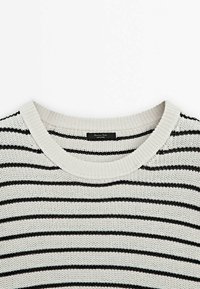 Pull blanc en maille avec des rayures horizontales noires et un col rond, avec une petite étiquette noire au niveau du col.