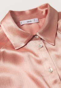 Gros plan d'une chemise en satin rose brillant avec un col pointu et des boutons blancs, montrant l'étiquette de la marque avec les informations de taille à l'intérieur du col.