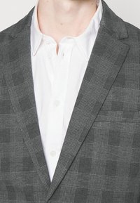 Blazer a quadre grigio realizzato in tessuto testurizzato, con rever a punte e una tasca sul petto, indossato sopra una camicia bianca con bottoni.