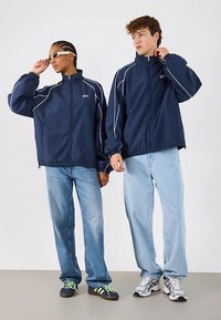 Tumsas zilas džemperis ar rāvējslēdzēju un baltu apdari, valkāts kopā ar gaiši zilām džinsu biksēm. Aksesuāri ietver saulesbrilles un dažādas apavu stilus.