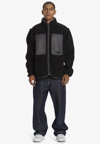 Svart fleecetröja med grå nylonframsida, hel dragkedja, stående krage och avslappnad passform, ihop med lösa mörka jeans och sneakers.