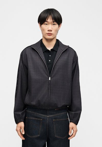 Bărbat tânăr purtând o jachetă închisă la culoare cu fermoar peste un tricou polo negru și blugi închiși la culoare, stând în picioare pe un fond deschis simplu.