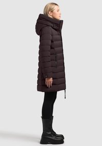 Ciemnofioletowa puchowa kurtka z wysokim kołnierzem, kieszeniami na suwak i gładką fakturą, w zestawieniu z czarnymi sznurowanymi butami wojskowymi i legginsami.