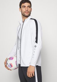 Witte sportjack met zwarte zijstrepen, ritsluiting en opvallende details op de mouwen. Model houdt een kleurrijk voetbal vast.