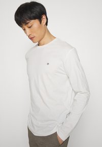 Jeune homme aux cheveux noirs portant une chemise blanche à manches longues et un pantalon à carreaux, les mains dans les poches, regardant vers le bas sur un fond blanc.