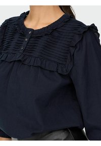 Blouse bleu marine avec un devant texturé et volanté, fermeture à boutons et encolure froncée. Fabriquée en tissu léger.