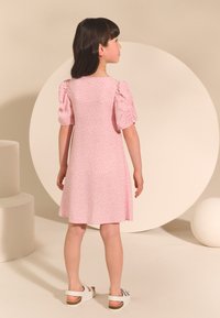 Ljusrosa klänning med kort puffärm, blommönster, utsvängd siluett och längd ovanför knäna. Bärs med vita sandaler. Bakre vy.