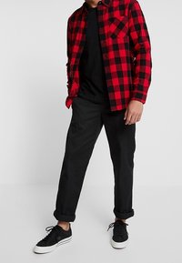 Camicia di flanella a scacchi rossi e neri, t-shirt nera sotto, pantaloni neri arrotolati e sneakers nere. Outfit casual con texture sovrapposte.