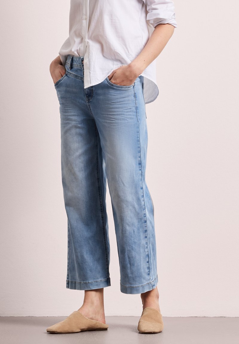 Persona vistiendo unos jeans de pierna ancha azul claro, camisa blanca de botones con las mangas remangadas y zapatos beige sin cordones, de pie con las manos en los bolsillos.