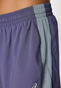 Bermudas deportivas de color púrpura oscuro con un panel de malla gris claro, cintura elástica y costuras finas; cuenta con un logo en el lado inferior.