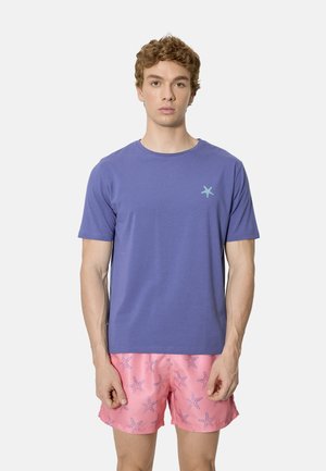 Joven con una camiseta lisa morada con un pequeño logotipo de estrella de mar y pantalones cortos rosas con un estampado de estrellas de mar azules, posando frente a un fondo blanco.