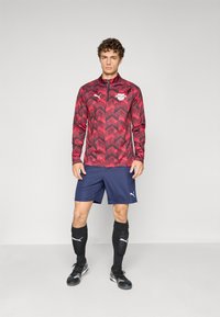 Puma treningsantrekk sett som inkluderer en rød og svart mønstret langermet topp, marineblå shorts, svarte sokker og fotballsko.