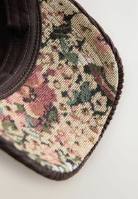 Brun hatt med en mönstrad brätte som har en texturerad blommönster i rosa, grönt och svart på en vävd beige bakgrund.