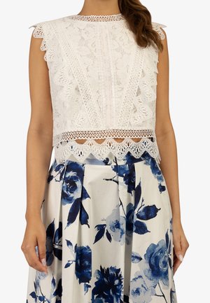 Crop top sans manches en dentelle blanche avec bords festonnés, assorti à une jupe blanche ornée de grandes impressions florales bleues et de plis.