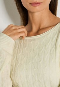 Eine Frau, die einen cremefarbenen, strukturierten Pullover trägt und den Ausschnitt hält, wobei ihr unteres Gesicht und der Hals sichtbar sind.