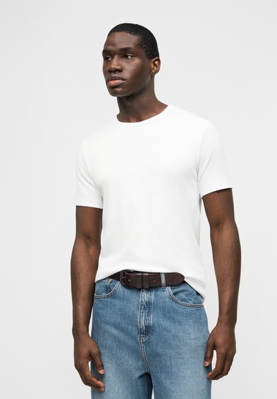 Άνδρας που φοράει λευκό κοντομάνικο μπλουζάκι με στρογγυλή λαιμόκοψη, το οποίο είναι φουσκωμένο μέσα σε μπλε τζιν με σκούρη καφέ ζώνη, στέκεται μπροστά σε απλό φόντο.