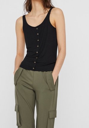 Kvinde iført en ærmeløs sort knapbelagt tanktop og olivengrønne cargobukser med hænderne i lommerne mod en ensfarvet baggrund.