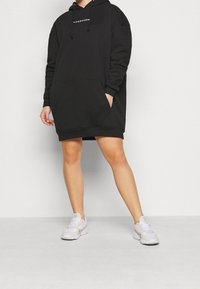 Černé oversized mikinové šaty s přední kapsou, kapucí se stahovací šňůrkou a žebrovanými manžetami, doplněné o bílé tenisky na neutrálním pozadí.