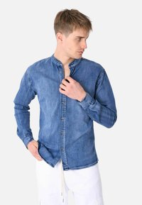 Camicia di jeans di un blu medio con colletto a fascia, maniche lunghe e chiusura anteriore con bottoni. Presenta una texture liscia e un fit casual.