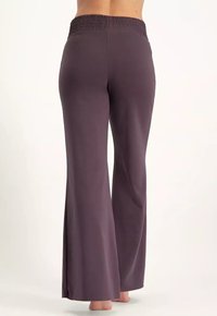 Kvinna som bär högmidjade, vida mörkpärletta byxor, står barfota på en ljus bakgrund, visar en bakvy från midjan till fötterna.