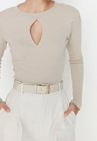 Haut beige à manches longues côtelé avec un détail goutte d'eau au niveau de l'encolure, porté avec une ceinture de couleur claire à boucle dorée et un pantalon blanc plissé.