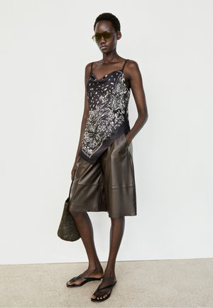 Donna con occhiali da sole, top nero fantasia, pantaloncini marroni al ginocchio, sandali neri e borsa intrecciata in mano, in piedi contro un muro bianco.