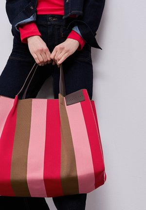 Henkilö, joka pukeutunut tummansiniseen farkkuasuun ja punaiseen yläosaan, pitää suuren vaaleanpunaisessa, punaisessa ja ruskeassa raidoitetussa kangaskassissa ruskeita kahvoja valkoista taustaa vasten.