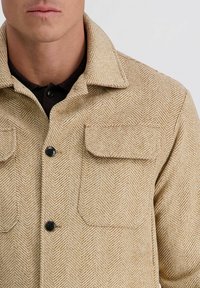 Close-up van een beige jasje met visgraatpatroon, uitgerust met twee klepzakken en zwarte knopen, gedragen over een zwarte polo.