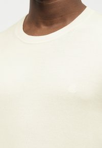 Κοντινό πλάνο ενός ατόμου που φοράει πουλόβερ πλεχτό εκρού με ριμπ λαιμόκοψη και διακριτικό ανάγλυφο λογότυπο στο στήθος.